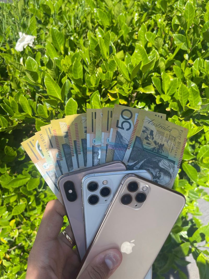 A hand holding used phones with Australian banknotes behind them, in front of a hedge